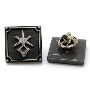 May include: Two square, dark gray metal pins with a stylized starburst and diamond design. One pin is shown with the pin back visible. The pins have a decorative border and rounded corners.