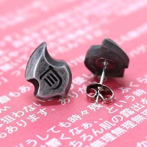 May include: A pair of silver-toned stud earrings. Each earring has a flame-like shape with a stylized design. The earrings are displayed on a pink background with Japanese text.