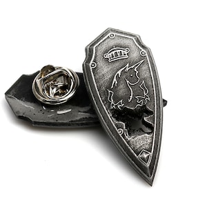 May include: A silver shield-shaped pin with a unicorn and crown design. The pin has a safety clasp on the back.