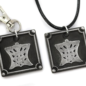 Puede incluir: Dos colgantes plateados con fondo negro y dise&ntilde;o plateado. Un colgante est&aacute; unido a un collar de cord&oacute;n negro y el otro a un llavero plateado.