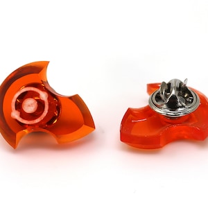 Op de afbeelding: Twee oranje plastic speldjes in de vorm van een halve maan. Het speldje links heeft een witte cirkel in het midden. Het speldje rechts heeft een zilveren achterkant.