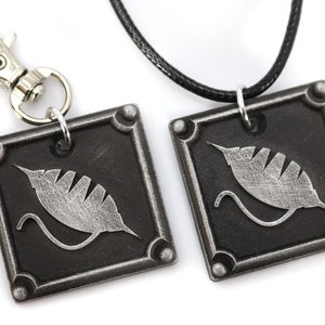 May include: Two silver-toned square pendants with a stylized leaf design. One pendant is attached to a black cord necklace, the other to a silver key chain.
