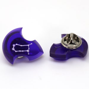 Op de afbeelding: Een paarse acryl pin met een wit Tweelingen sterrenbeeld ontwerp. De pin heeft een zilveren metalen sluiting aan de achterkant.