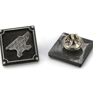 May include: A silver and black metal pin with a square shape. The pin features a silver anvil design on a black background. The back of the pin has a metal clasp.