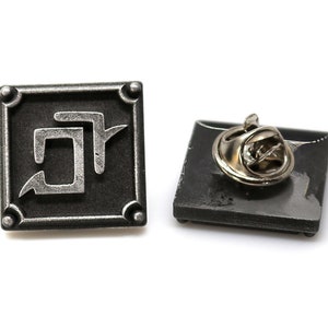 May include: A silver and black metal pin with a stylized symbol on a black background. The pin has a safety clasp on the back.