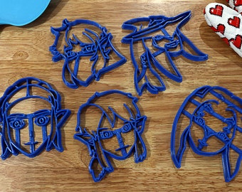 May include: Five blue plastic cookie cutters shaped like cartoon characters with elf ears and large eyes. The cutters are on a wooden surface.