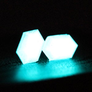 K&ouml;nnte beinhalten: Zwei leuchtende, sechseckige Formen in Teal auf schwarzem Hintergrund.