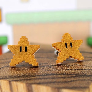 May include: A pair of gold star-shaped earrings with black eyes. The earrings are made of a glittery material and are inspired by the classic video game character.
