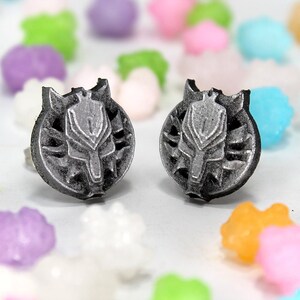 Op de afbeelding: Een paar zilverkleurige oorbel studs met een wolfskop ontwerp. De oorbellen zijn geplaatst tegen een witte achtergrond met kleurrijk snoep.