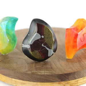 May include: Three resin earbud tips in different colors and patterns. The tips are shaped like a teardrop and have a swirl pattern. The colors are green, black, and orange.