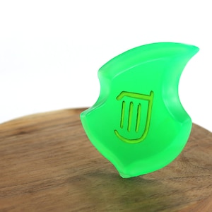 May include: A green, translucent, leaf-shaped charm with a harp symbol in the centre. The charm is sitting on a wooden surface.