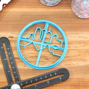May include: A blue plastic cookie cutter shaped like a city skyline. The cutter is round and has a detailed design of buildings and a river.