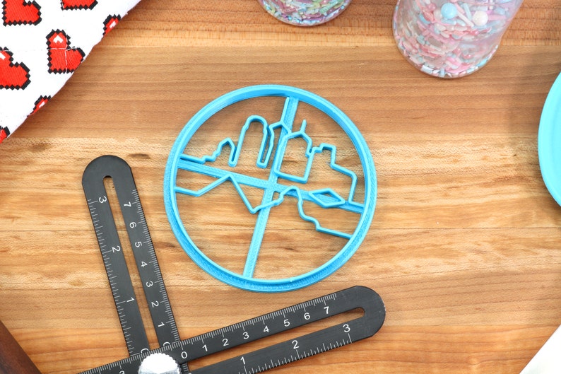 May include: A blue plastic cookie cutter shaped like a city skyline. The cutter is round and has a detailed design of buildings and a river.