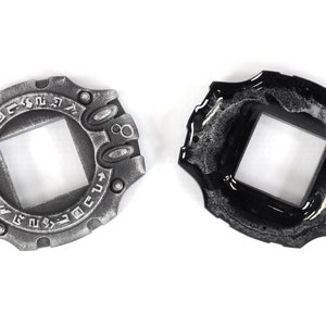 May include: Two silver and black metal Digimon Digivices with a square window in the center. The Digivices have a circular design with raised edges and a series of symbols around the edge.