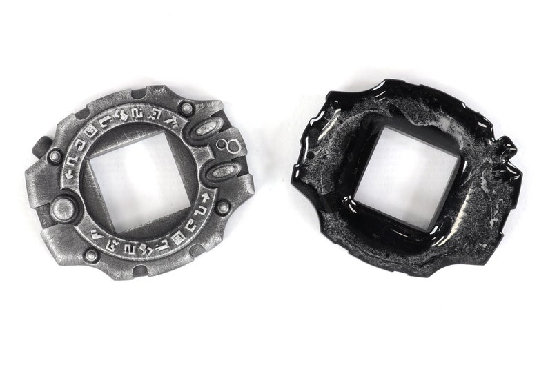 May include: Two silver and black metal Digimon Digivices with a square window in the center. The Digivices have a circular design with raised edges and a series of symbols around the edge.