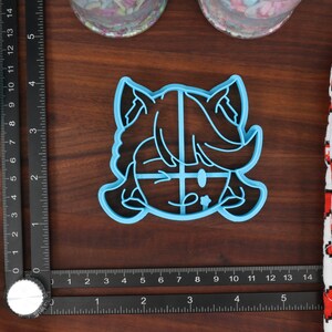 May include: A blue plastic cookie cutter shaped like a cartoon character with a surprised expression. The character has large eyes, a small mouth, and a large head with short hair. The cookie cutter is on a wooden surface.