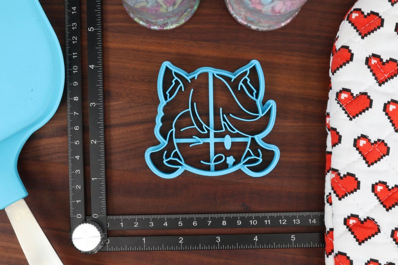 May include: A blue plastic cookie cutter shaped like a cartoon character with a surprised expression. The character has large eyes, a small mouth, and a large head with short hair. The cookie cutter is on a wooden surface.