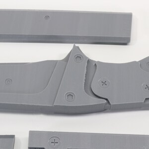 May include: A gray 3D printed model of a knife with a detailed design. The knife has a textured handle and a sharp blade. The model is made up of multiple pieces that are joined together with screws.