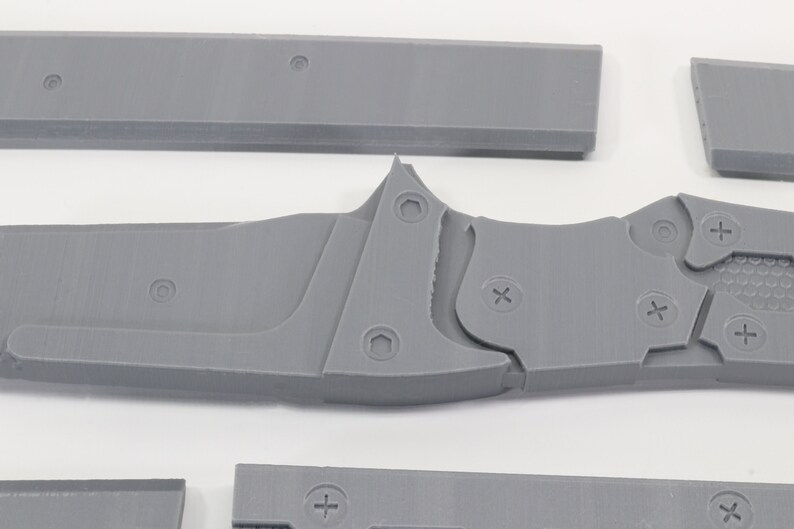 May include: A gray 3D printed model of a knife with a detailed design. The knife has a textured handle and a sharp blade. The model is made up of multiple pieces that are joined together with screws.