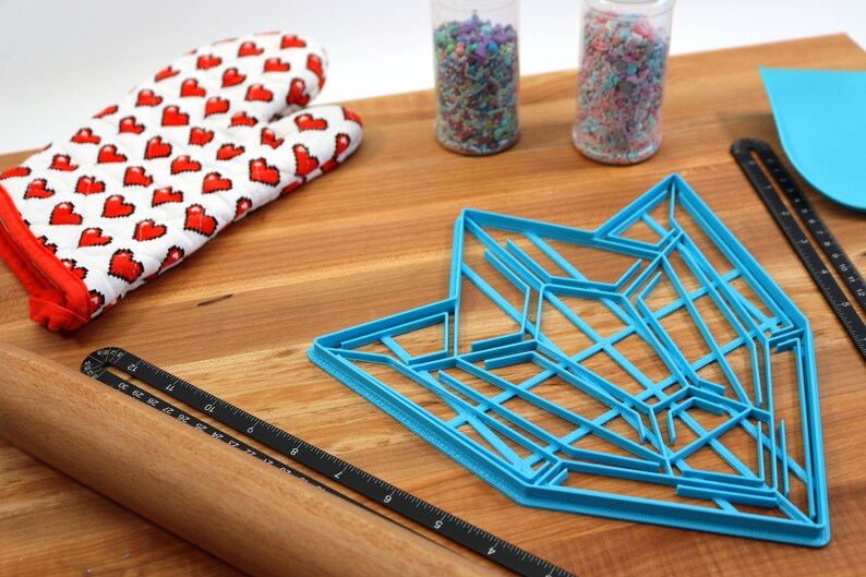 Puede incluir: Un cortador de galletas de pl&aacute;stico azul en forma de estrella geom&eacute;trica, un rodillo de madera, un guante de horno rojo y blanco y dos frascos de chispas en una superficie de madera.