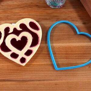 May include: A heart-shaped cookie with a red jelly filling and a blue heart-shaped cookie cutter on a wooden surface.