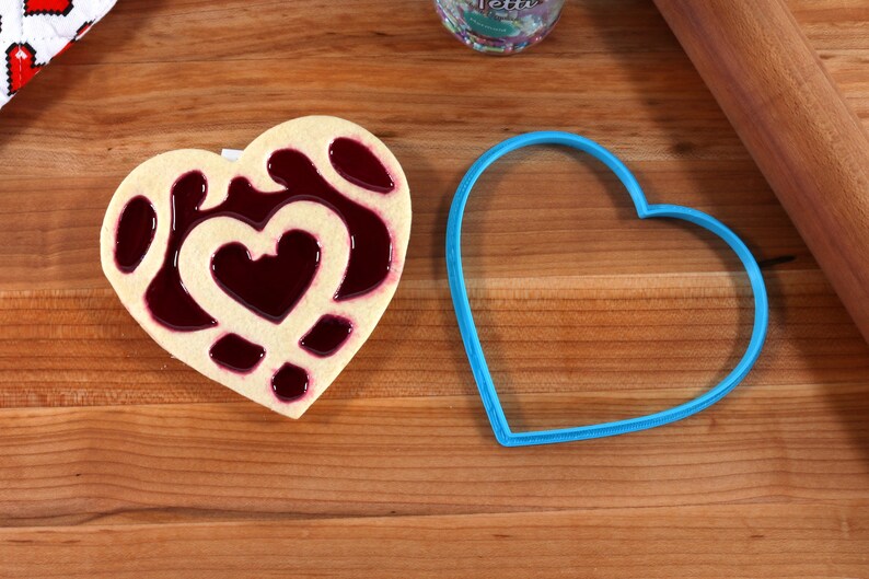 May include: A heart-shaped cookie with a red jelly filling and a blue heart-shaped cookie cutter on a wooden surface.