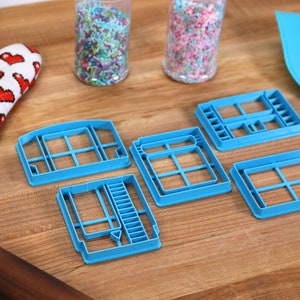 May include: Five blue plastic cookie cutters shaped like video game cartridges on a wooden surface. Two jars of colorful sprinkles are in the background.
