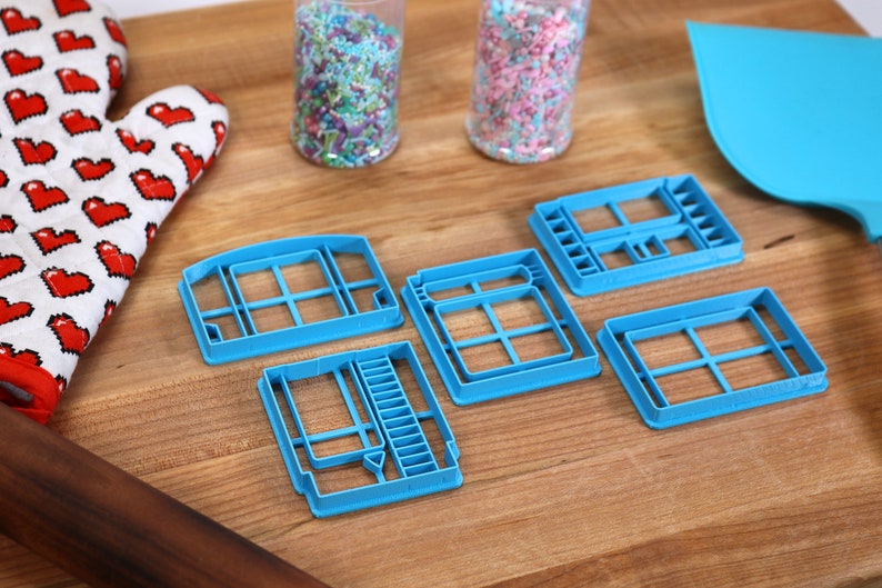 May include: Five blue plastic cookie cutters shaped like video game cartridges on a wooden surface. Two jars of colorful sprinkles are in the background.