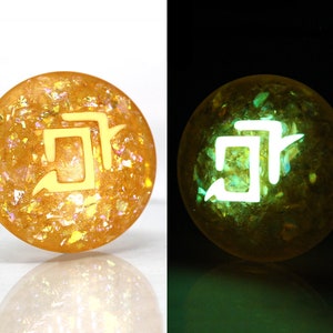 May include: A round, yellow resin cabochon with a white symbol and a glittery, iridescent finish. The cabochon glows green in the dark.
