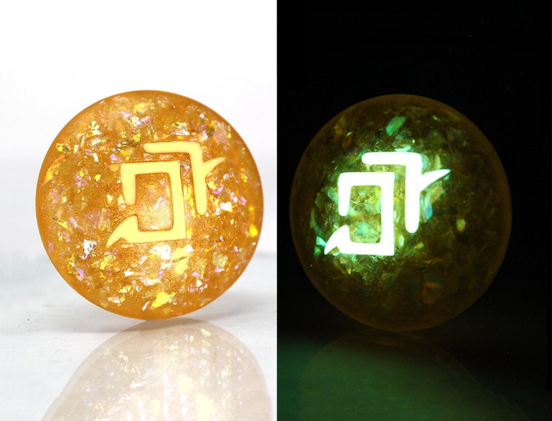 May include: A round, yellow resin cabochon with a white symbol and a glittery, iridescent finish. The cabochon glows green in the dark.