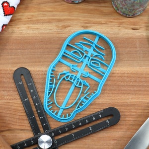 May include: A blue plastic cookie cutter shaped like a person's face with an open mouth. The cookie cutter is on a wooden surface.