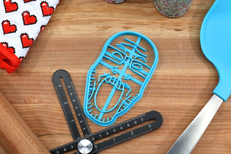 May include: A blue plastic cookie cutter shaped like a person's face with an open mouth. The cookie cutter is on a wooden surface.