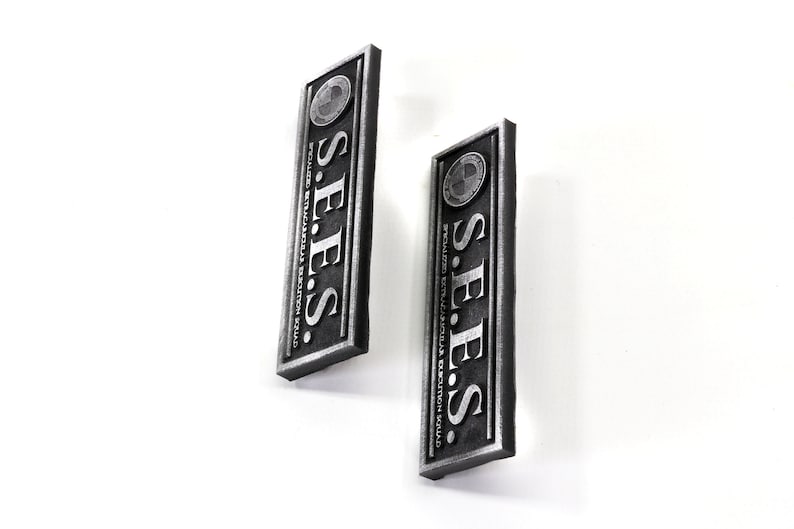 May include: Two silver metal rectangular signs with the text "S.E.E.S." and "SECURE EXTRATERRESTRIAL EXTRACTION SQUAD" in black lettering.