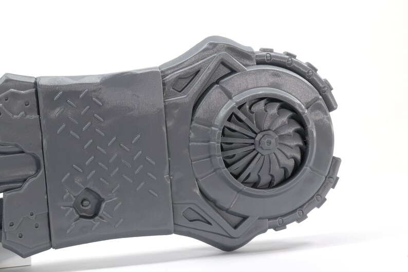 May include: A gray 3D printed model of a futuristic weapon with a detailed design. The weapon has a large, circular fan blade on one end and a textured surface on the other.