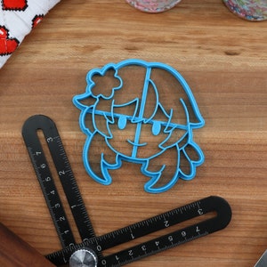 May include: Blue plastic cookie cutter shaped like a cartoon character with a flower in their hair. The character has a smiling face and long hair.