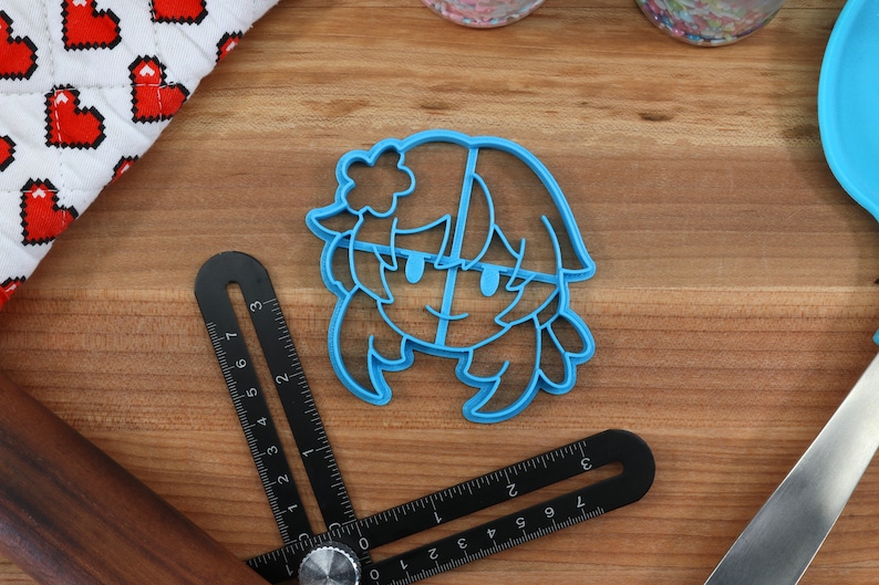 May include: Blue plastic cookie cutter shaped like a cartoon character with a flower in their hair. The character has a smiling face and long hair.