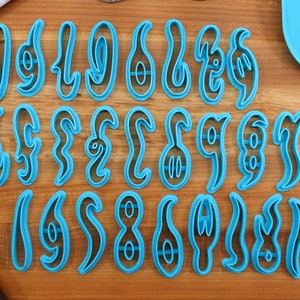 May include: A set of 24 blue plastic cookie cutters shaped like different letters of the alphabet. The cutters are arranged in three rows of eight on a wooden surface.