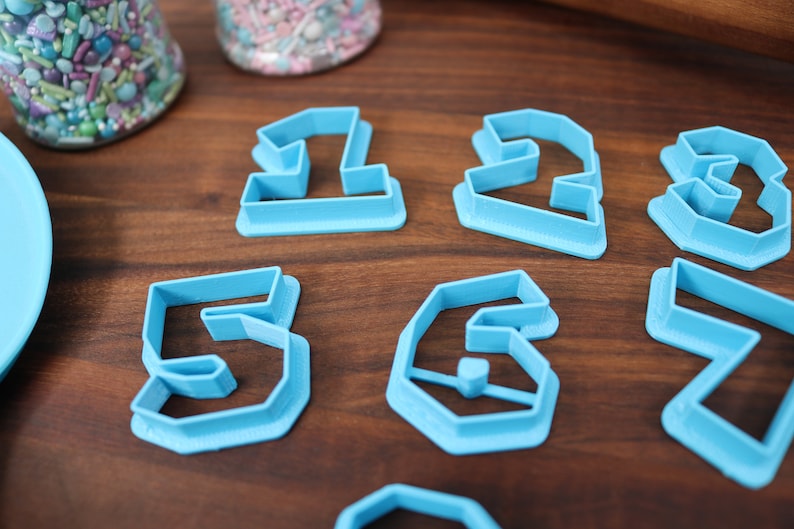 May include: A set of blue plastic cookie cutters shaped like the numbers 1 through 7. The cutters are on a wooden surface.