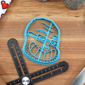 May include: A blue plastic cookie cutter shaped like a person wearing glasses and smiling. The cookie cutter is on a wooden surface.
