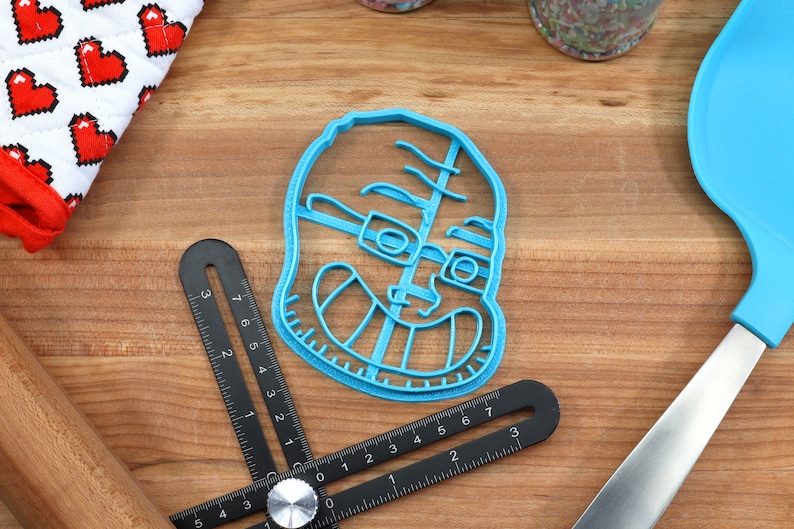 May include: A blue plastic cookie cutter shaped like a person wearing glasses and smiling. The cookie cutter is on a wooden surface.
