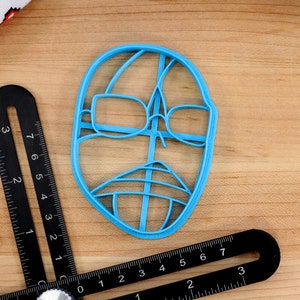 May include: A blue plastic cookie cutter shaped like a face with glasses and a mustache.