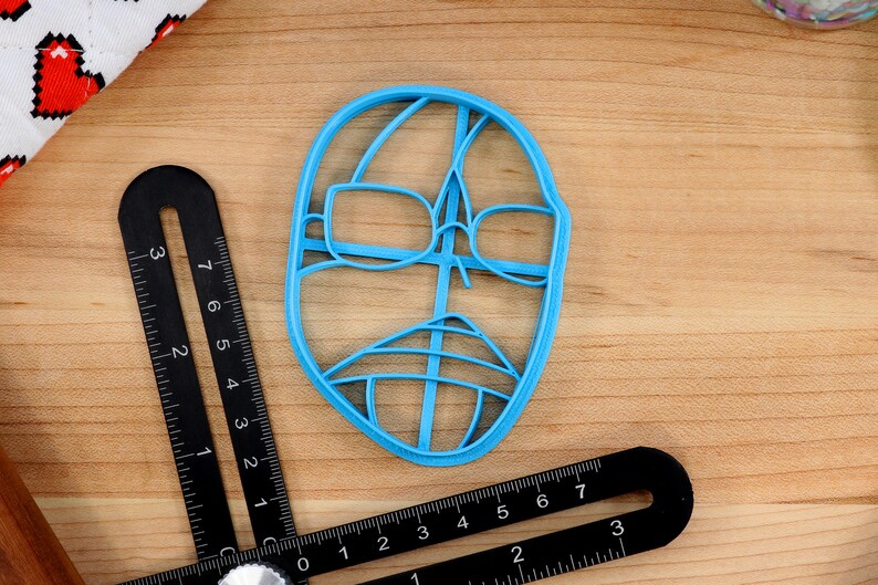May include: A blue plastic cookie cutter shaped like a face with glasses and a mustache.