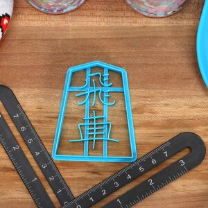 May include: A light blue plastic cookie cutter with Japanese characters. The cutter is on a wooden surface, next to a black ruler, a blue spatula, and a white oven mitt with red pixelated hearts.