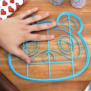 May include: A blue plastic cookie cutter shaped like a cartoon animal face. The cutter is on a wooden surface and is large enough to fit a hand inside.