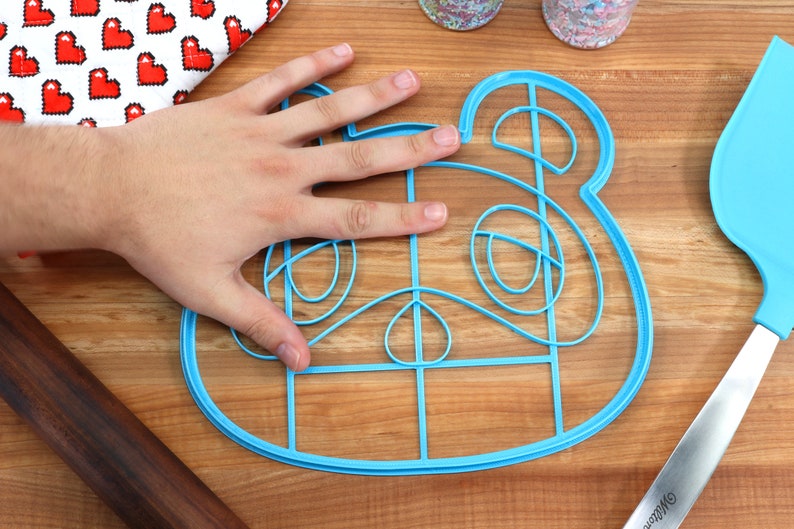 May include: A blue plastic cookie cutter shaped like a cartoon animal face. The cutter is on a wooden surface and is large enough to fit a hand inside.