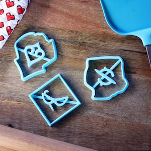 May include: Three blue plastic cookie cutters on a wooden surface. The cutters are shaped like a heart with a dollar sign inside, a diamond with a dollar sign with a line through it, and a square with a pair of glasses inside.