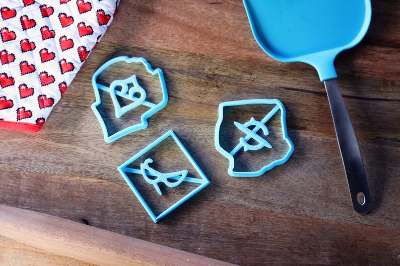 May include: Three blue plastic cookie cutters on a wooden surface. The cutters are shaped like a heart with a dollar sign inside, a diamond with a dollar sign with a line through it, and a square with a pair of glasses inside.