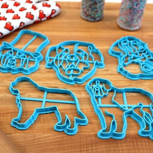 May include: A collection of five light blue dog-shaped cookie cutters, including a dog wearing a hat, a dog's head, and two full-body dog shapes. A red and white heart-patterned oven mitt, sprinkles, a blue spatula, and a rolling pin are also visible.