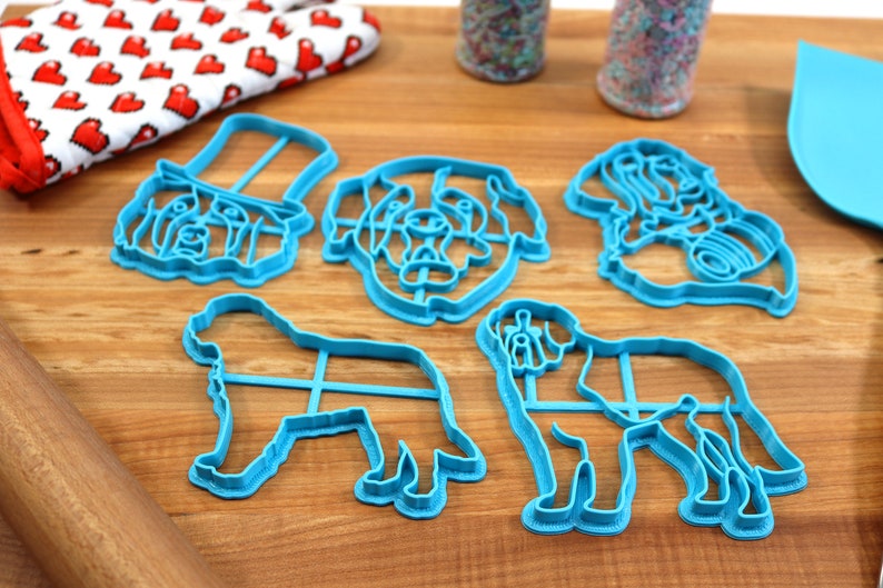 May include: A collection of five light blue dog-shaped cookie cutters, including a dog wearing a hat, a dog's head, and two full-body dog shapes. A red and white heart-patterned oven mitt, sprinkles, a blue spatula, and a rolling pin are also visible.