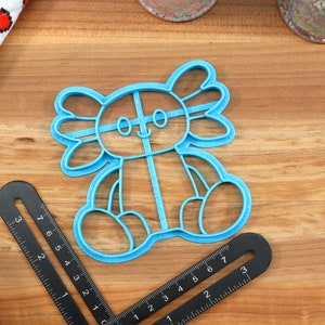May include: A blue plastic cookie cutter shaped like an axolotl. The axolotl is sitting with its legs crossed and has a happy expression on its face.